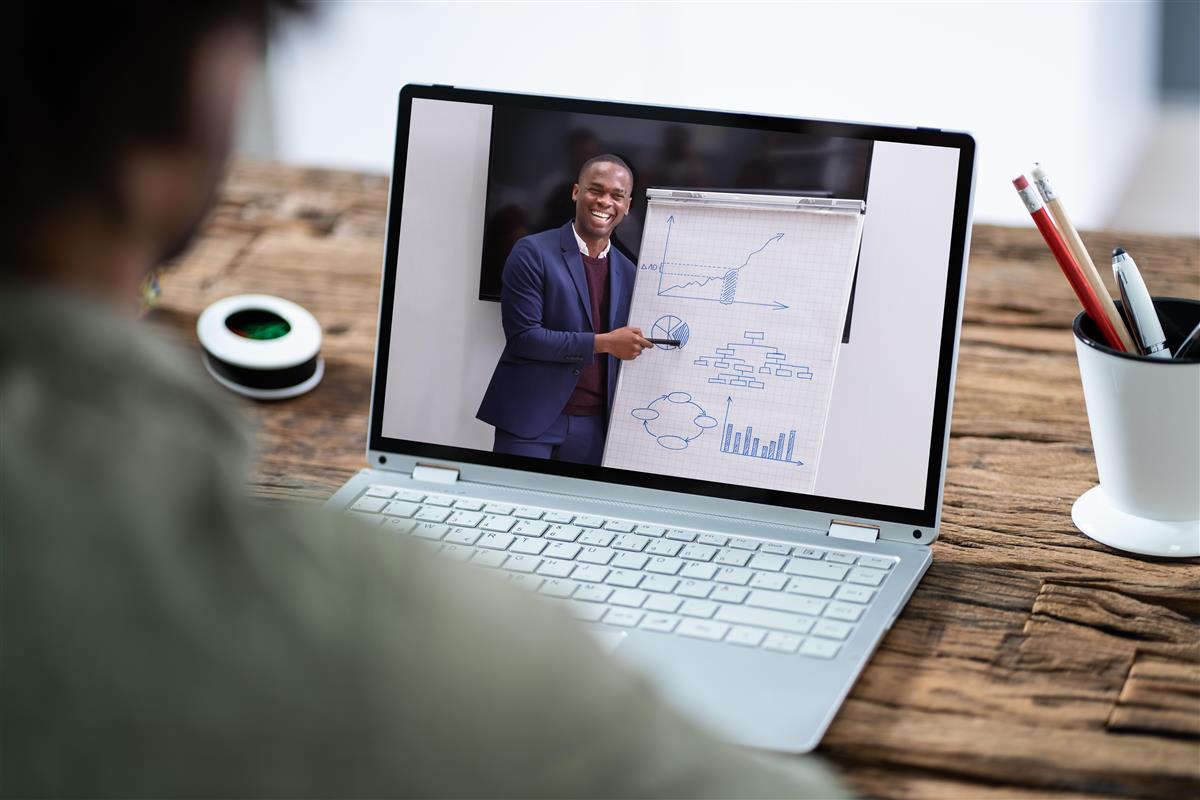 Bilde av person som ser på en dataskjerm der en mann står foran en flipover med ulike diagrammer - Klikk for stort bilde