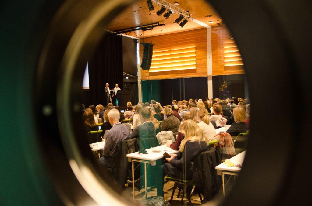 Bilde tatt gjennom et rundt vindu inn til konferanselokalet der deltakerne sitter i skoleoppsett - Klikk for stort bilde