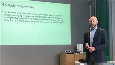 Olsen holder presentasjon, står foran skjerm med en slide om brukermedvirkning - Klikk for stort bilde